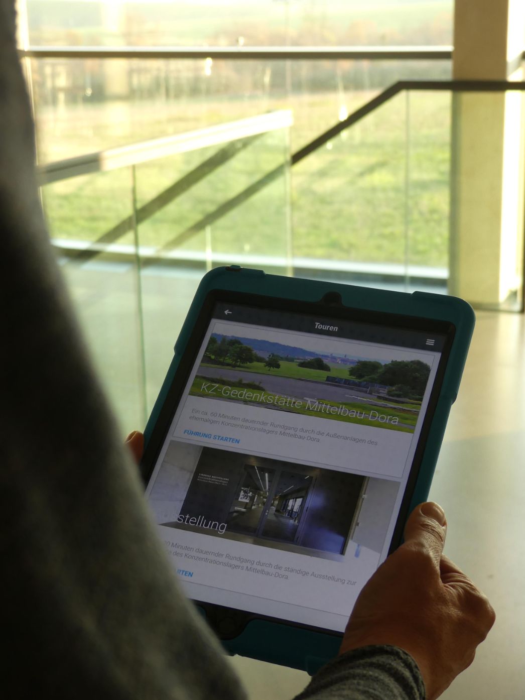 A person holds a tablet in their hand. On the screen you can see the open app of the permanent exhibition of the Mittelbau-Dora Concentration Camp Memorial.
