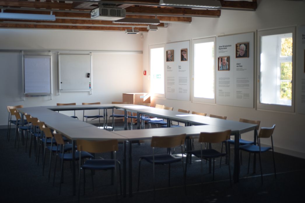 A seminar room at the Mittelbau-Dora concentration camp memorial. In the room, there are tables arranged in a rectangle. Chairs are placed around them. Light falls into the room through 3 windows. A beamer hangs under the ceiling. 