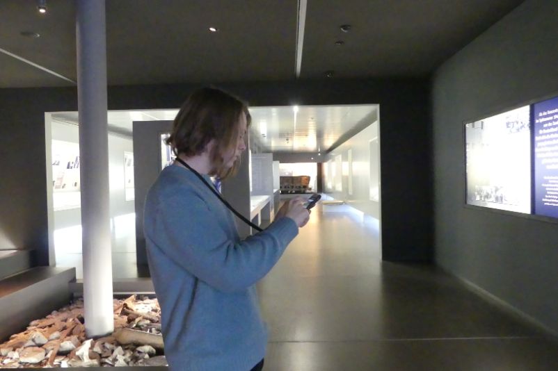 Ein junger Mann im Eingangsbereich der Dauerausstellung, der ein Handy in einer schwarzen Hülle an einem schwarzen Band um den Hals trägt. Er tippt gerade auf den Bildschirm. 