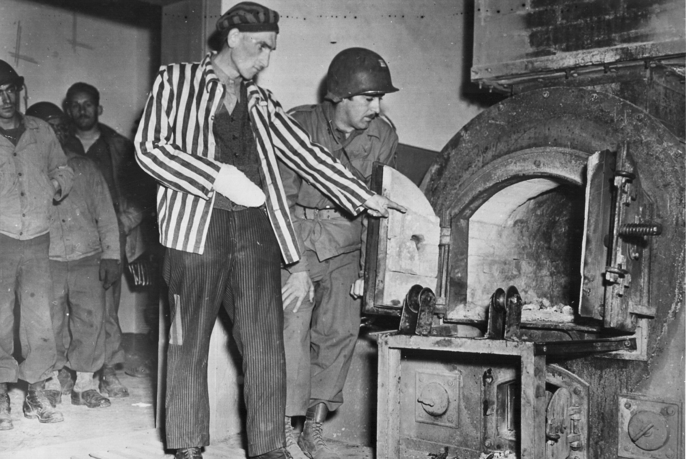 Im Zentrum des Bildes ist ein Mann in Häftlingskleidung, der neben einem US-Soldaten steht und in einen der Krematoriumsoefen zeigt. Im Hintergrund stehen weitere US-Soldaten und sehen durch einen Türbogen zu.
