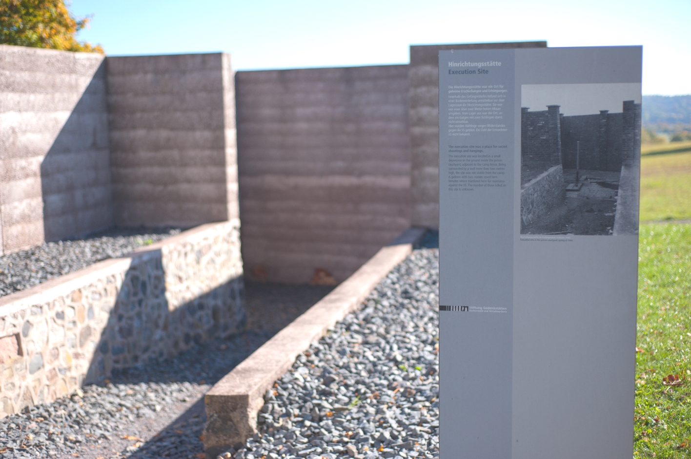 Im Vordergrund ist auf der rechten Seite die Informationstafel an der Hinrichtungsstätte zu sehen, auf der ein historisches Vergleichsbild zur heutigen Gestaltung der Hinrichtungsstätte angeboten wird. Im Hintergrund ist eine Vertiefung mit hohen Betonmauern zu sehen, die die ehemalige Hinrichtungsstätte nachbildet.