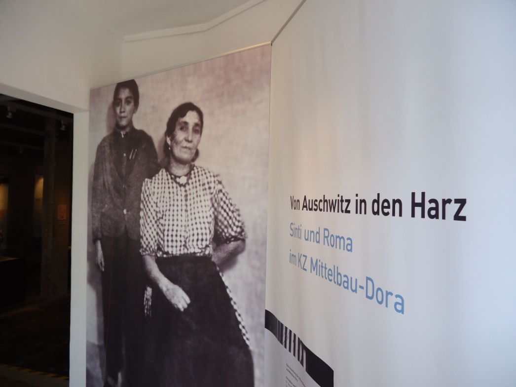 Links ist ein Türrahmen zu sehen. Daneben stehen vor einer weißen Wand zwei Banner. Das linke zeigt ein Schwarz-Weiß-Foto von einem Jungen und einer Frau. Auf dem rechten steht der Titel der Ausstellung.
