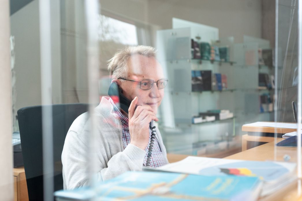 Ein Mitarbeiter der Information hat einen Telefonhörer in der Hand und blickt nach vorne durch eine Plexiglasscheibe, die die Information vom Foyer trennt.