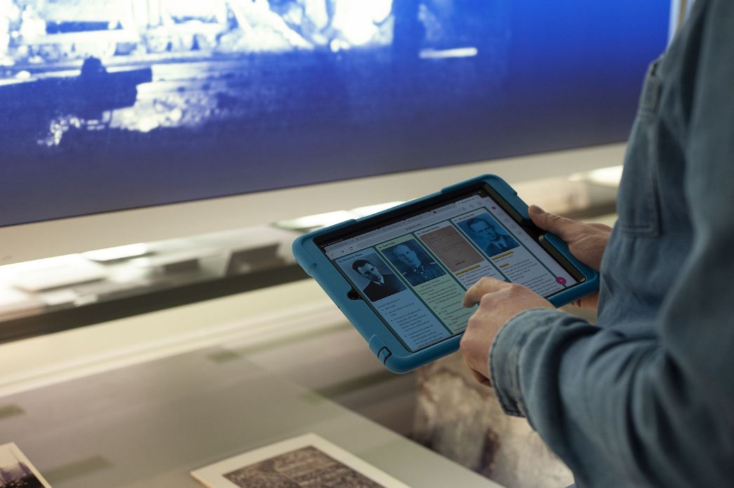 The photo shows the hands of a person using a tablet to access biographies. 