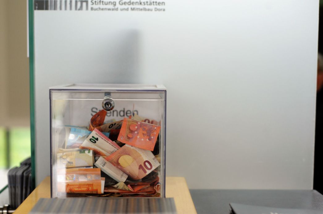The picture shows a transparent donation box filled with all kinds of euro bills.