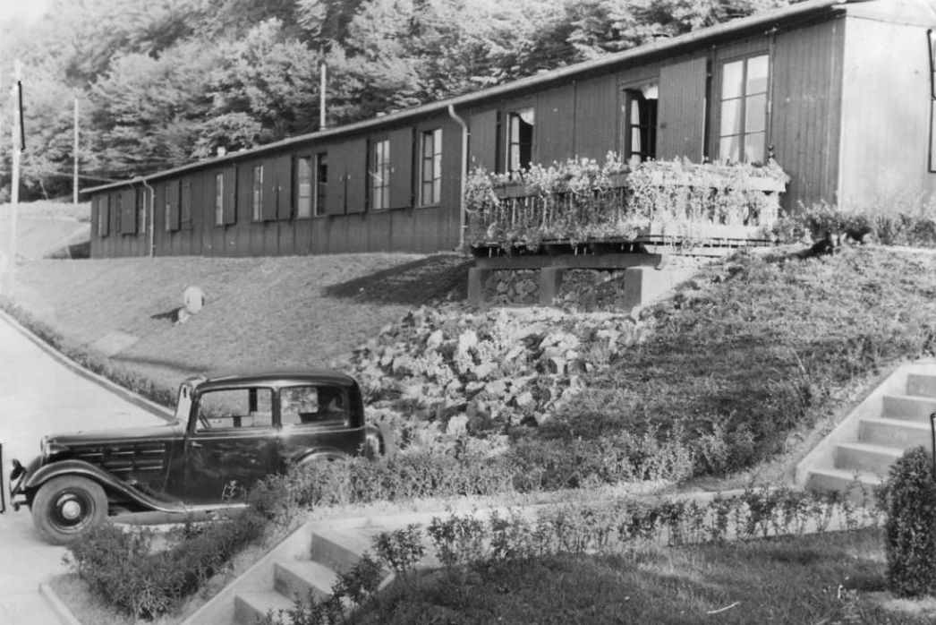 Blick auf eine der Unterkunftsbaracken, mit Balkon und gepflegter Gartenanlage, im SS-Bereich des Konzentrationslagers Mittelbau-Dora. Unterhalb des Balkon parkt ein Auto.