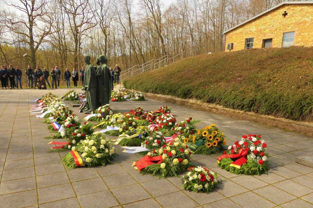 Several wreaths of flowers with colored bows are placed in front of and next to a group of figures. The crematorium building can be seen on the right. Participants of a memorial event dressed in black stand in the background.