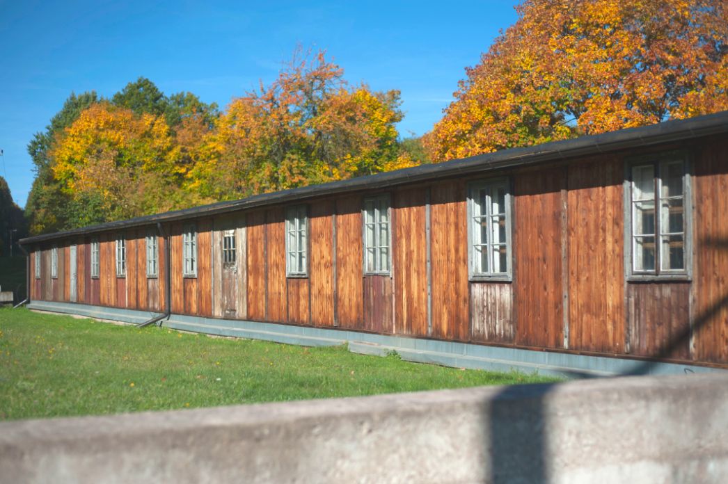The photo shows the wooden facade of the carpentry barracks. Behind the barracks there are deciduous trees that look autumnal.