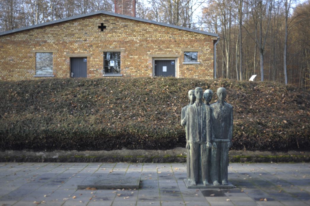 Ein Platz mit einer Statue und Gedenkplatten. Im Hintergrund sieht man das ehemalige Krematorium.