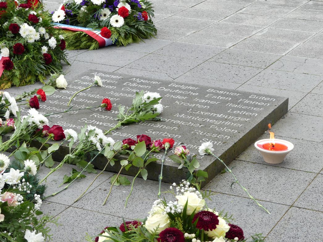 Eine Gedenkplatte auf dem Boden. Auf und neben der Gedenkplatte liegen Blumen. Eine brennende Kerze steht neben der Gedenkplatte.