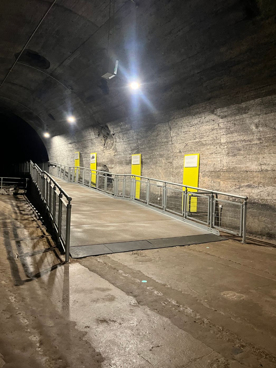 Ein Stollen unter der Erde, einige Lampen beleuchten ihn. Eine Rampe führt weiter in den Stollen hinein.