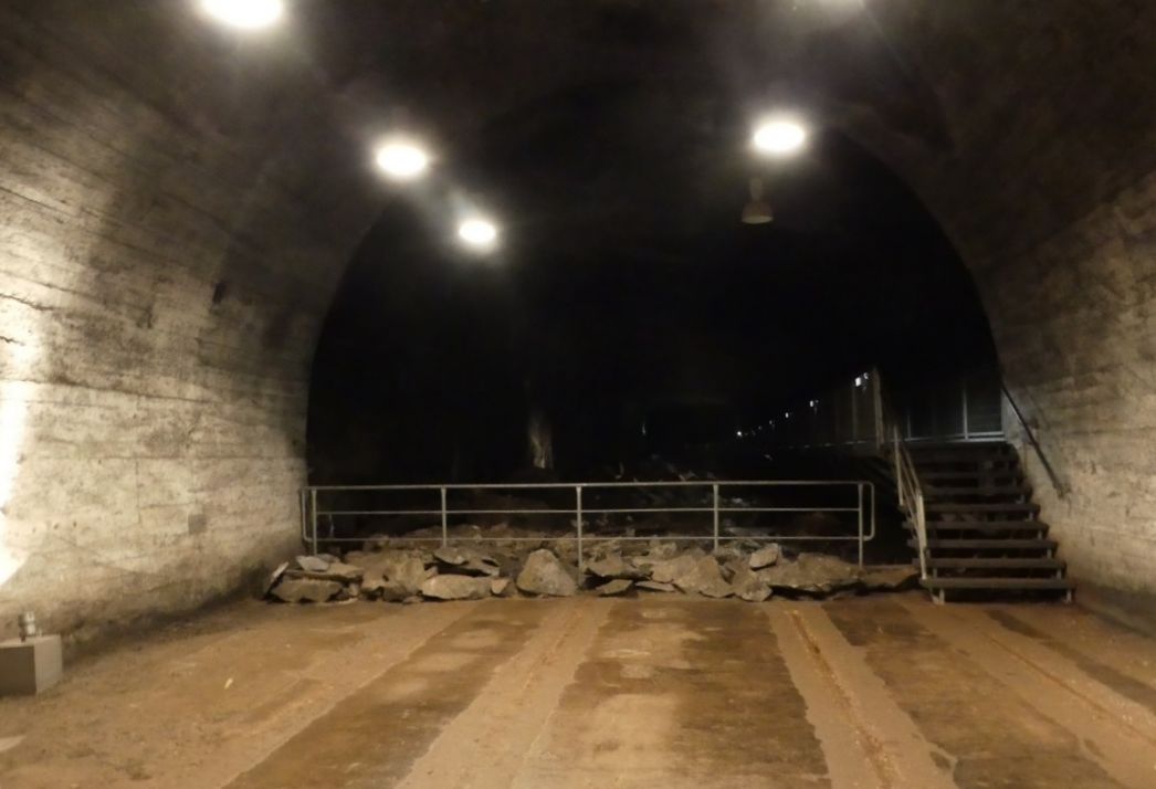 A gallery underground, some lamps illuminate it from above. A metal staircase leads to a narrow platform. Underneath, large chunks of stone. 