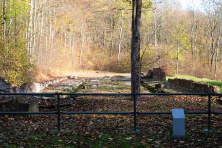 A metal railing can be seen, which was set up in front of a depression in which the remains of the foundation can be seen. In front of the railing there is a small information board that reveals that it is the remains of the former laundry.