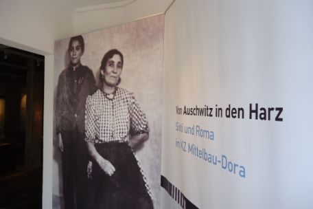Links ist ein Türrahmen zu sehen. Daneben stehen vor einer weißen Wand zwei Banner. Das linke zeigt ein Schwarz-Weiß-Foto von einem Jungen und einer Frau. Auf dem rechten steht der Titel der Ausstellung.