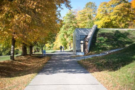 Path made of concrete slabs under trees. The exhibition building is built into a kind of embankment. 