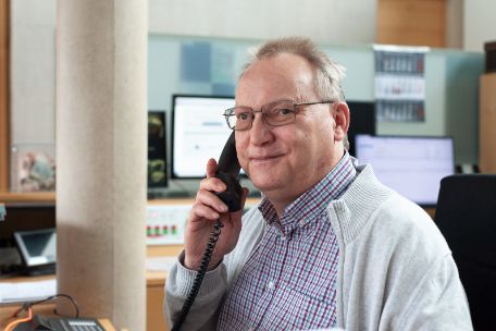 Ein Mitarbeiter der Information hat einen Telefonhörer in der Hand und lächelt in die Kamera.