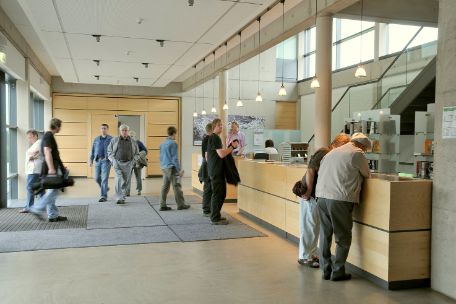 Ein Foyer mit einem Tresen. Besucher:innen stehen davor und sprechen mit Mitarbeiter:innen.