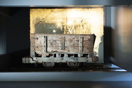 Das Foto zeigt einen Grubenhunt in der Ausstellung. Obwohl das Exponat gut ausgeleuchtet ist, wirkt die Umgebung eher dunkel. Im Hintergrund ist eine wandfüllende Fotografie des Stolleninneren angebracht.