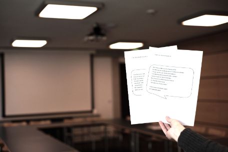 A hand holding up two worksheets with testimonies of former prisoners in easy language. In the background, the tables and blackboard of a seminar room can be seen.