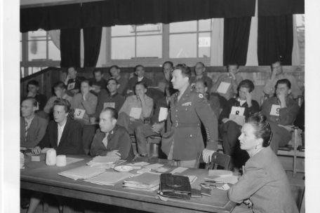 The defenders are sitting at a table in the foreground. One of them is standing. In the background, the defendants sit in two rows on chairs, with numbers around their necks.