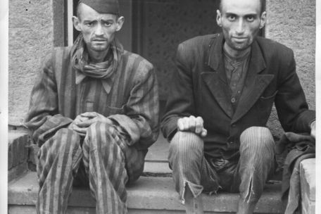Zwei abgemagerte befreite Häftlinge sitzen nebeneinander auf Treppenstufen und blicken direkt in die Kamera.