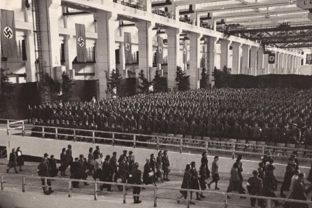 Die Raketenmontage-Halle F 1 in Peenemünde, die sich durch ihre Größe und Höhe auszeichnet. Im Vordergrund ziehen Menschen in die Halle ein. Dahinter sind Menschen zu erkennen, die zum Appell angetreten sind. Nahe der Seitenwand der Halle sind mehrere Hakenkreuzbanner angebracht.