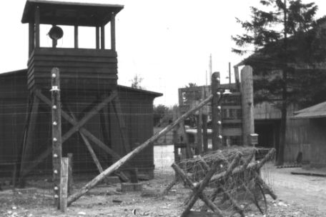 Zu sehen ist ein Wachturm aus Holz neben einem halboffen stehenden hölzernen Lagertor. Davor ist ein Spanischer Reiter mit Stacheldraht zu sehen, der neben dem Zugangsweg steht.