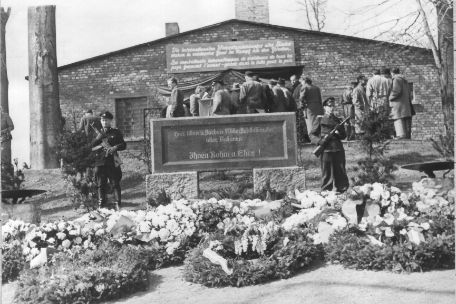  In the foreground the memorial stone with the inscription: "Here suffered and died resistance fighters of all nations. To them glory and honor." To the right and left of it two NVA soldiers with rifles. In the background the building of the crematorium. Participants of the memorial ceremony are standing in front of it.
