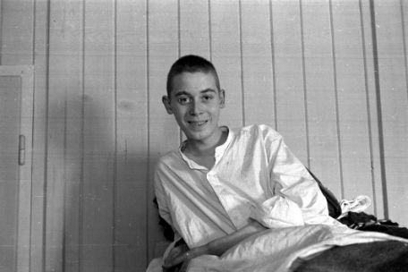 A young man with a shaved head is seen sitting on a hospital bed and smiling at the camera.