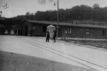 Im Vordergrund stehen zwei Personen auf der Lagerstrasse und unterhalten sich. Dahinter ist die Baracke der Politischen Abteilung neben dem Lagereingang zu sehen. Vor dem Tor stehen weitere Personen.