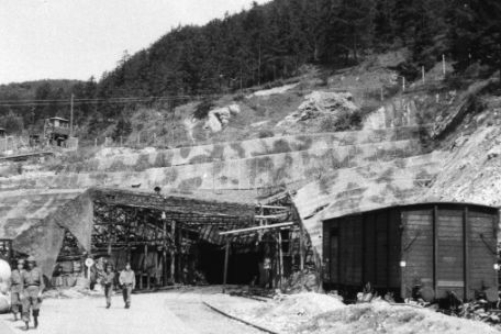 Blick auf den mit Tarnnetzen getarnten Eingang zum Fahrstollen A des Mittelwerks. Rechts ist ein Eisenbahnwaggon zu sehen. Links entfernen sich vier US-Soldaten, jeweils in Zweiergruppen, vom Stollen.