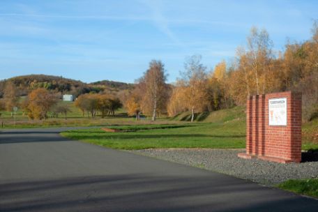 On the right of the picture is a small piece of wall made of red brick. Attached to this section of wall is a plaque with a map showing the routes of the death marches from Mittelbau-Dora concentration camp. The camp road leads past this section of the wall on the left.