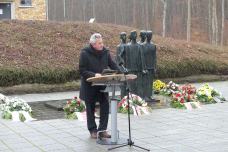 Prof. Dr. Wagner giving a speech in the memorial square in front of the crematorium. He is dressed in dark clothes and stands at a narrow lectern. In the background are wreaths of flowers arranged around the memorial plaque and group of figures of the memorial square.