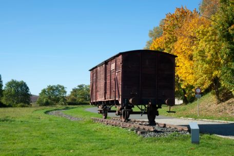The photo shows an old railway carriage of the German Reichsbahn in autumn. The wagon is standing on a small piece of track in a meadow. There is a small information board in front of the wagon.