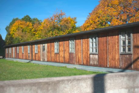 Das Foto zeigt die Holzfassade der Tischlereibaracke. Hinter der Baracke stehen herbstlich anmutende Laubbäume.