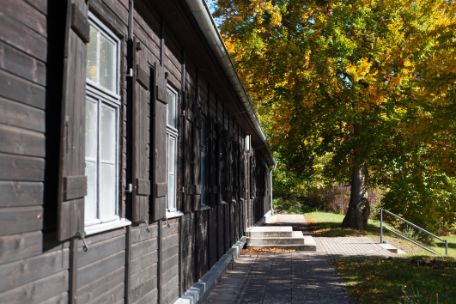 Das Foto zeigt die schwarze hölzerne Außenfassade der rekonstruierten Holzbaracke. Rechts ist erkennbar, dass sie an einem Hang erbaut wurde. Eine Treppe führt hinunter. Neben der Treppe steht ein Baum, dessen Blätter herbstlich verfärbt sind.