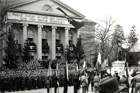 Truppen vor dem Theater, Denkmal rechts, am Theater Hakenkreuzflagge und das Spruchband „Adolf Hitler schafft den Weltfrieden – Gib ihm deine Stimme!“