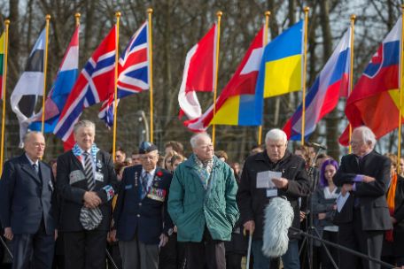 Several older men are standing in front of a crowd of people holding up flags. One has a piece of paper in his hand and is talking. One wears a uniform with insignia, one has a red angle and a number on his jacket. 