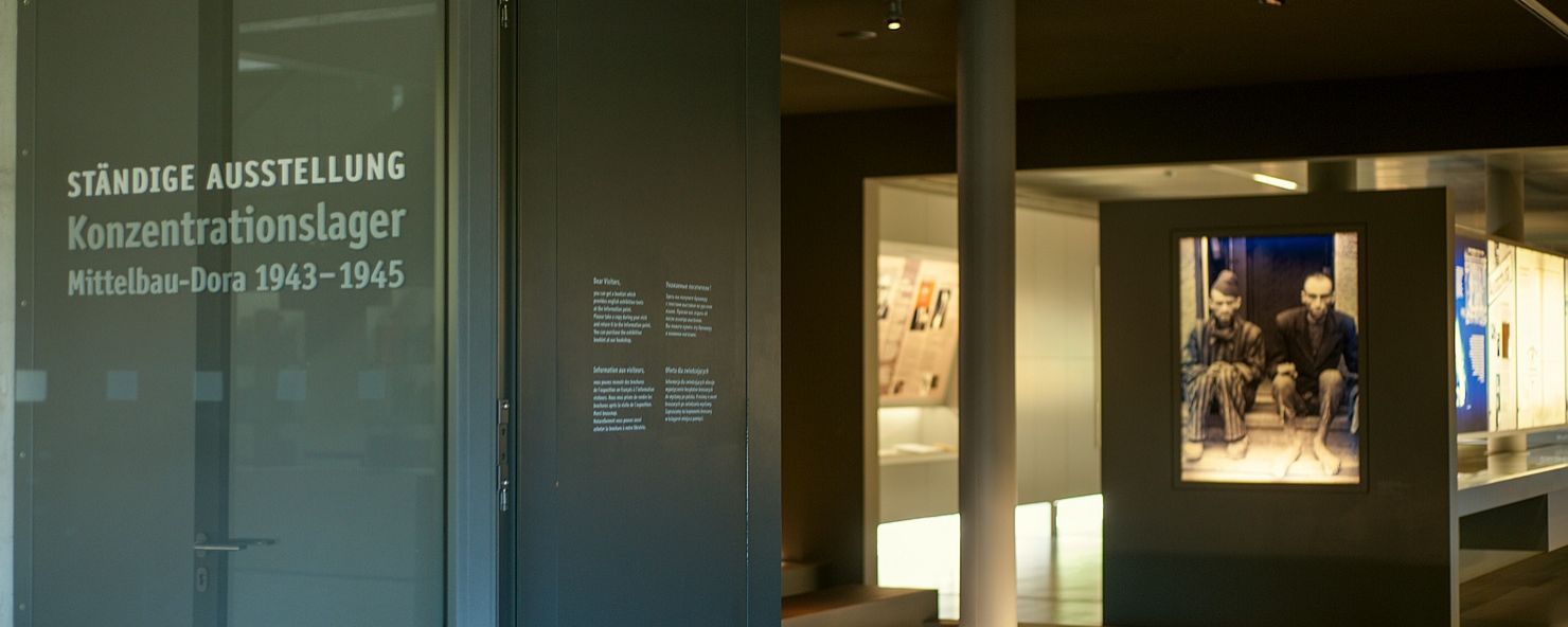 Blick durch den Eingang in die Dauerausstellung. Der Einführungsraum mit dem großen Bild zweier befreiter Häftlinge, die auf einer Stufe vor einer Tür sitzen. Im Vordergrund des Bildes links neben dem Eingang eine Glastür mit der Aufschrift "Ständige Ausstellung". 