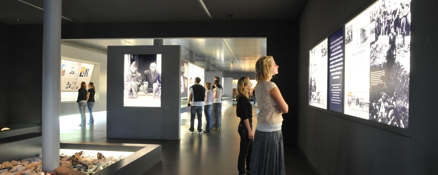 A dark room. On the walls, backlit panels with photos and texts. Visitors look at the panels. 