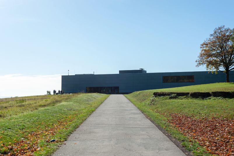 Das Bild zeigt den Zugangsweg zum Gedenkstättengebäude. Das Gebäude ist im Hintergrund zu sehen. Es handelt sich um einen mehrstöckigen Flachbau mit moderner Optik.