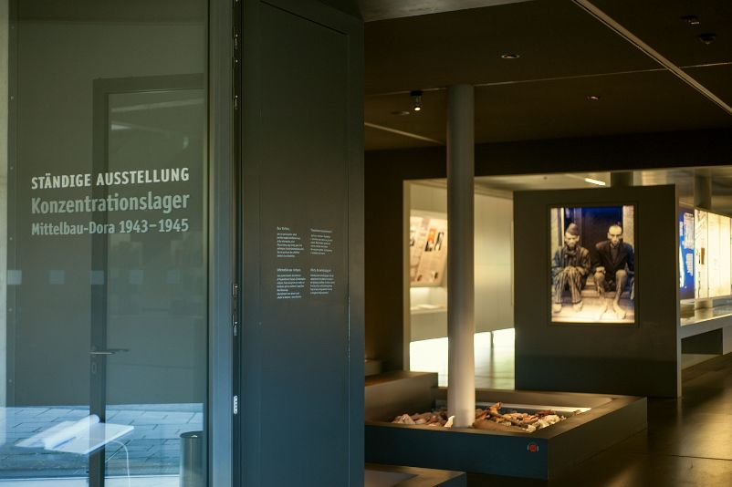 View through the entrance into the permanent exhibition. The introduction room with a large picture of two liberated prisoners sitting on a step in front of a door. In the foreground of the picture, to the left of the entrance, a glass door with the inscription "Permanent Exhibition". 