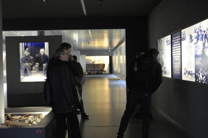 View into the dark exhibition room. The panels on the walls are illuminated from behind and show photos and texts. Visitors stand in the aisle between them. 