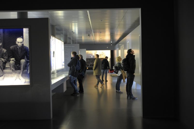 Zu sehen sind Bersucher:innen in einem der Austellungskorridore der Dauerausstellung, die rechts und links in die Inhalte vertieft sind, vereinzelt umhergehen oder sich unterhalten.