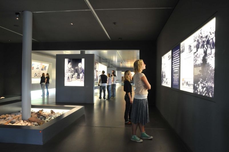 A dark room. On the walls, backlit panels with photos and texts. Visitors look at the panels. 