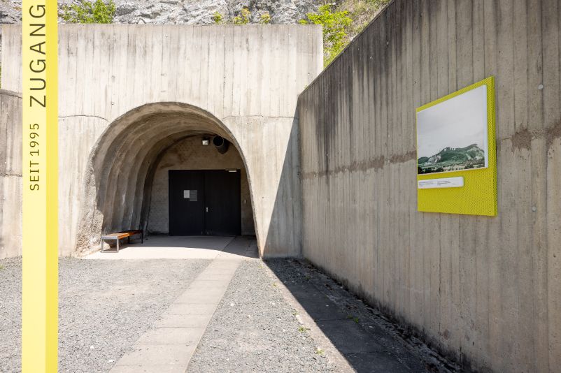 The photo shows the entrance to the new access tunnel from 1995. A yellow stele marking the entrance can be seen in the foreground on the left. On the wall to the right is a picture of the blasting of the tunnel system in 1947/48.