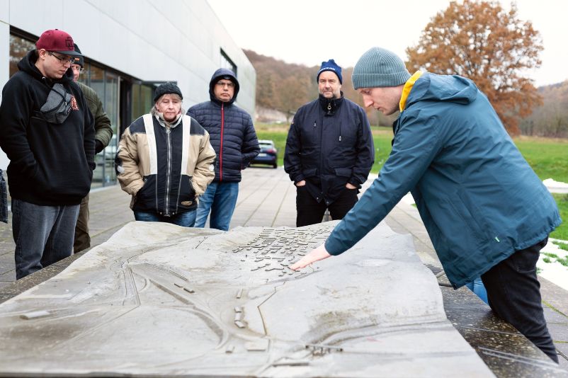 A small group stands around a model of the camp. A guide points to a spot and explains something. 