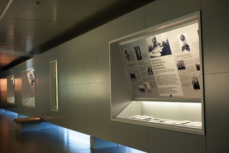 The photo shows an exhibition wall in the permanent exhibition. The section in focus deals with perpetrator biographies. At the bottom of the wall are showcases with exhibits, and above them are information panels on the perpetrators' biographies.