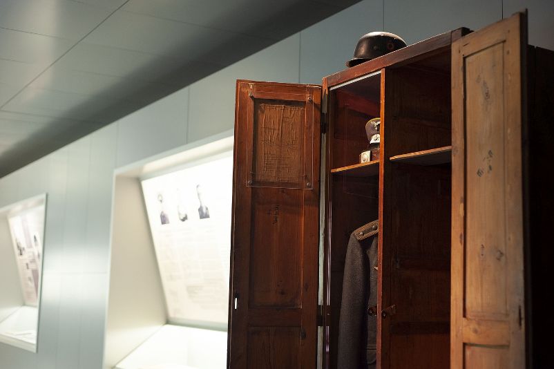 The photo shows an exhibit of a wooden locker of the Waffen-SS. In the locker you can see a Luftwaffe uniform and an umbrella cap. On top of the cabinet is a helmet.