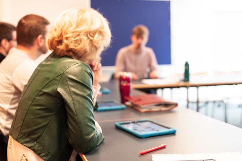 Das Foto zeigt eine Seminarsituation. Junge Menschen sitzen in kreisförmig angeordneten Tischen. Darauf Notizen und Tablets. 